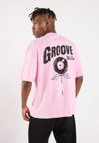 Pink short-sleeve t-shirt featuring a graphic design of a vinyl record and a hand holding a flower; printed text "Groove in Bloom" on back.