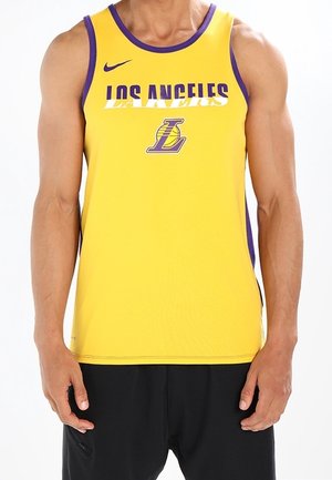 Homme portant un maillot de basketball jaune des Los Angeles Lakers avec des bordures violettes et un short noir, debout devant un fond blanc.
