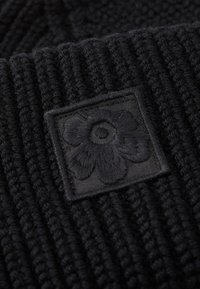 Schwarze gestrickte Mütze mit gerippter Textur und einem quadratischen schwarzen Patch, das ein geprägtes Blumendesign zeigt. Das Material wirkt weich und warm.