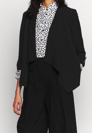 Kvinde iført en sort åben blazer over en hvid bluse med sorte polkaprikker, kombineret med sorte bukser med høj talje.
