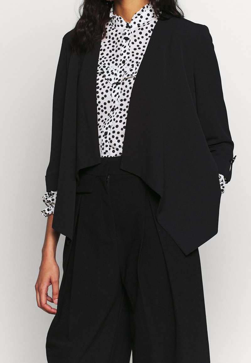 Femme portant un blazer noir ouvert sur un chemisier blanc à pois noirs, associé à un pantalon noir taille haute.