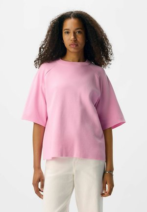 Pink oversized kortærmet skjorte med rund halsudskæring, lavet af blødt stof. Båret med hvide bukser, der har en afslappet pasform.