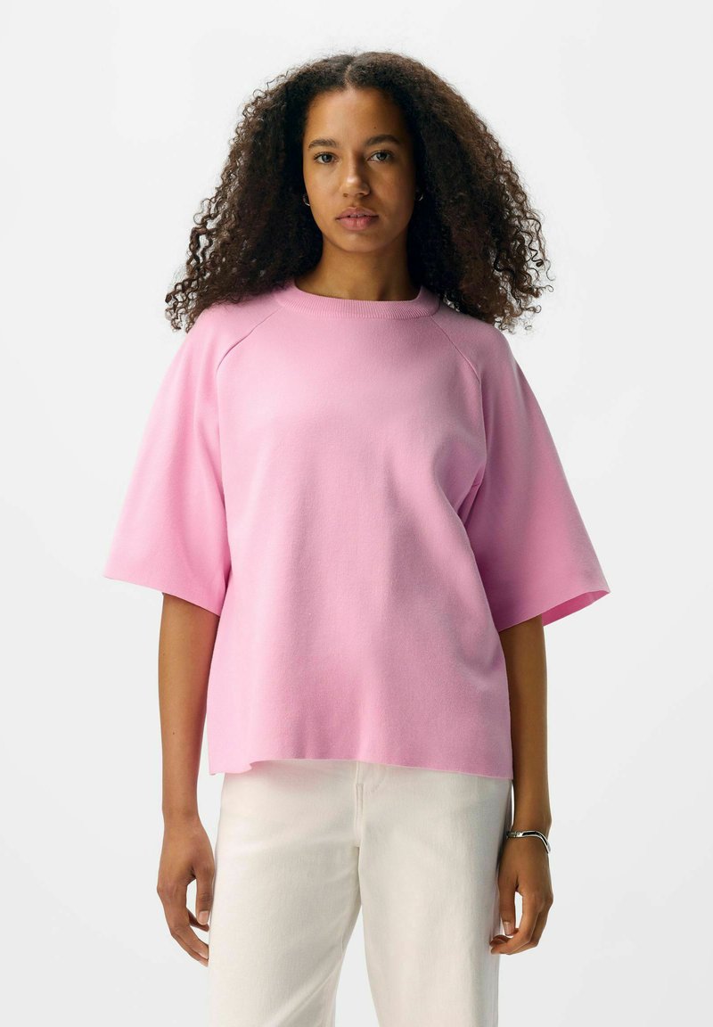 Pink oversized kortærmet skjorte med rund halsudskæring, lavet af blødt stof. Båret med hvide bukser, der har en afslappet pasform.
