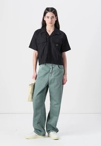 Camisa preta cropped com botões, equipada com dois bolsos no peito, combinada com calças de ganga verde folgadas. A modelo segura uma bolsa bege clara, vestindo ténis brancos.