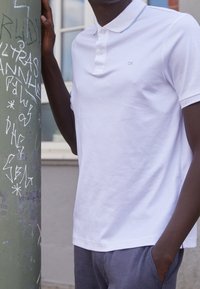 Vit pikétröja med en treknappsknut, ljusblå detaljer på kragen och en subtil "CK"-logotyp. Mjuk, len bomullstextur.