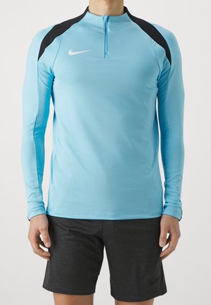 Mann trägt ein hellblaues, langärmliges Sportoberteil mit schwarzen Schulterakzenten und schwarze Shorts vor einfachem Hintergrund.