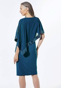 Turquoise jurk met een vloeiende cape, versierd met pailletten langs de randen. Gladde textuur, kniehoogte en een aansluitend ontwerp.