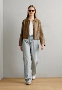 Rjava usnjena kratka jakna, bela srajca, svetlo modri visokotlačni kavbojke in srebrni teniski. Model nosi temne sončne očala, svetla ozadja.