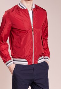 Uomo che indossa una giacca bomber rossa con zip, polsini e cintura a coste a strisce, pantaloni blu e mani in tasca su uno sfondo neutro.