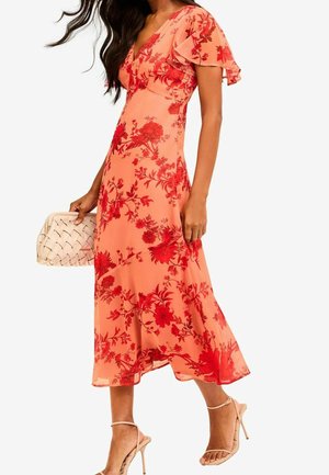Vrouw draagt een perzikkleurige midi-jurk met rode bloemenpatronen, zwierige mouwen, houdt een beige geweven clutch vast en beige sandalen met hoge hakken.