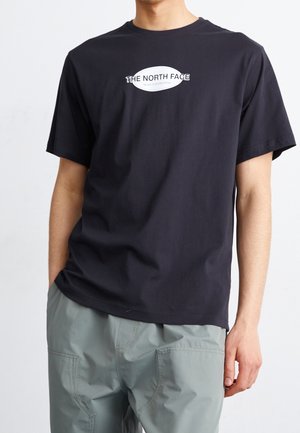 Persoon draagt een zwart T-shirt met korte mouwen met het logo "THE NORTH FACE" en grijze broek, staand tegen een effen achtergrond.