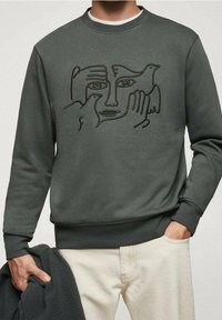 Bluza z okrągłym dekoltem z abstrakcyjną grafiką liniową przedstawiającą twarz i dwa ptaki, noszona przez osobę trzymającą ciemną kurtkę i ubraną w jasne spodnie.