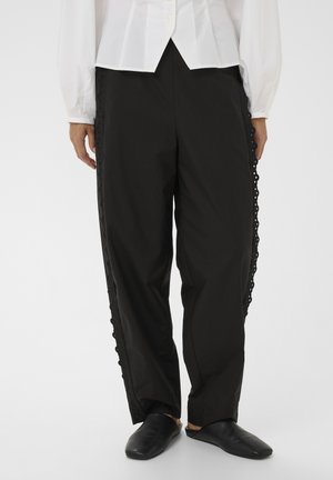 Vrouw draagt zwarte broek met brede pijpen en oogjeskant aan de zijkanten, witte blouse met knopen en zwarte instapschoenen, op een effen achtergrond.