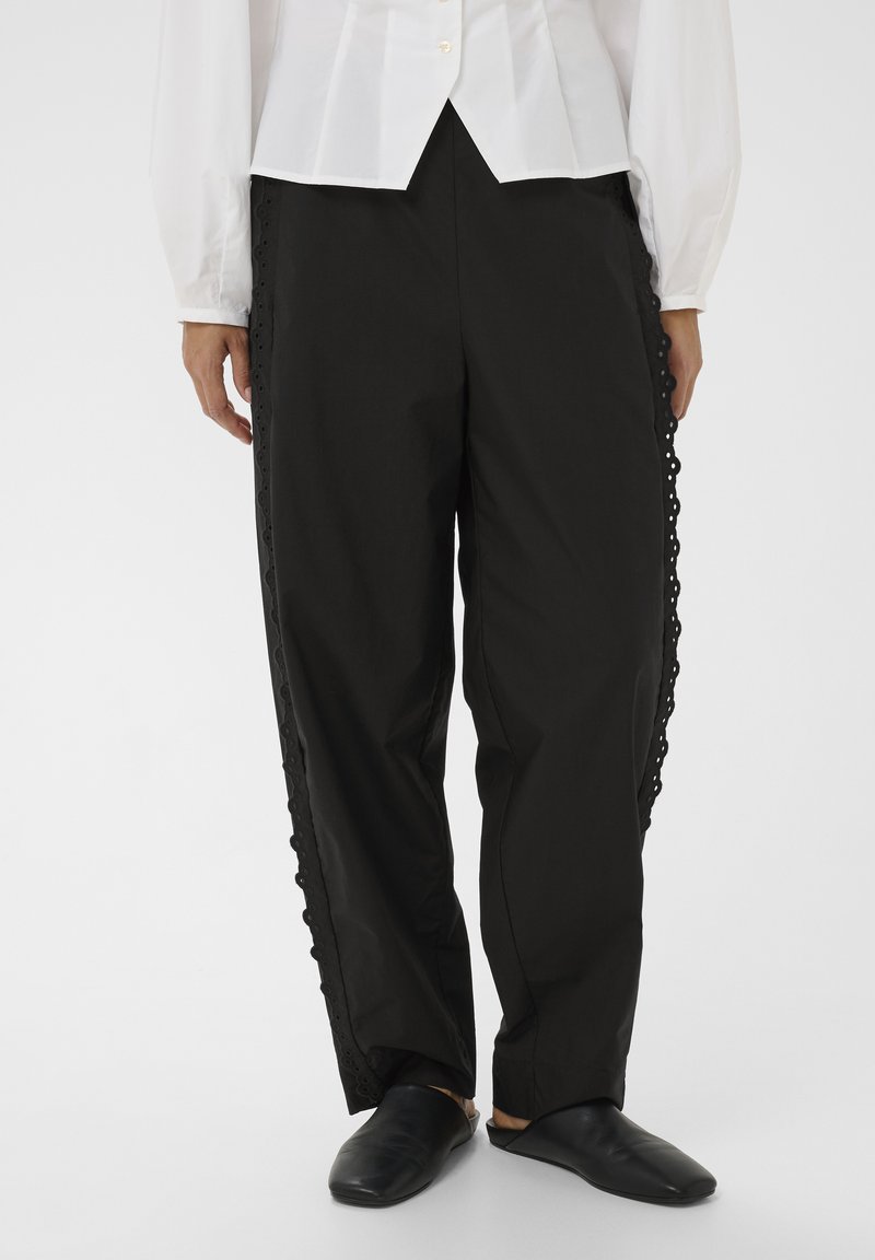 Femme portant un pantalon noir à jambes larges avec bordure œillet sur les côtés, un chemisier blanc boutonné et des chaussures noires sans lacets sur fond uni.