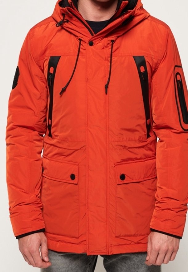 Doudoune orange avec capuche, dotée de poches zippées noires, d'un cordon de serrage ajustable et de poignets côtelés. Texture lisse et design spacieux.