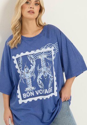 Blonde Frau trägt ein lockeres blaues T-Shirt mit Hummern und dem Aufdruck "BON VOYAGE" auf der Vorderseite, kombiniert mit hellblauen Jeans.