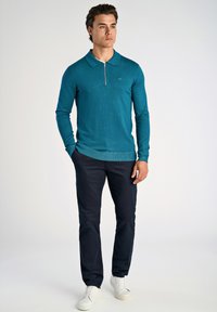 Teal långärmad pikétröja med dragkedja vid kragen, stickad textur och logotyp på bröstet, kombinerad med mörkblå byxor och vita sneakers.