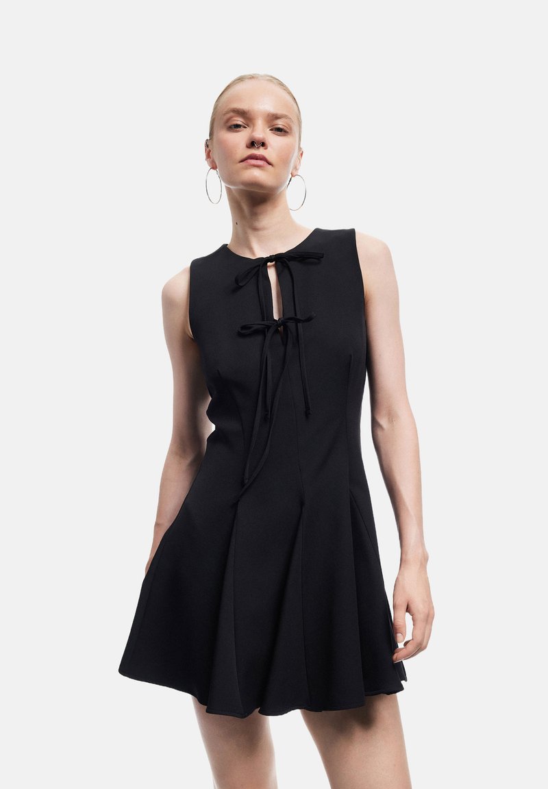Sort langærmet kjole med rund halsudskæring, plisseret skirt og sløjfe detalje foran. Glat stof med en let struktureret pasform.