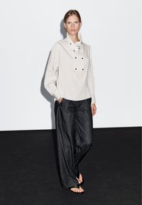 Světle šedá dlouhá košile s pruhy a vysokým límcem, s černými knoflíkovými detaily, v kombinaci s tmavými širokými džínovými kalhotami a sandály.