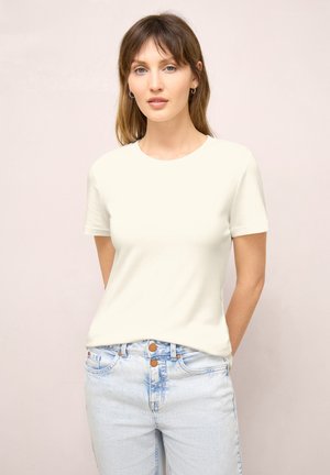 Frau mit hellbraunen Haaren, die kleine Creolen, ein einfaches weißes Kurzarmshirt und hellblaue, hoch taillierte Jeans trägt.