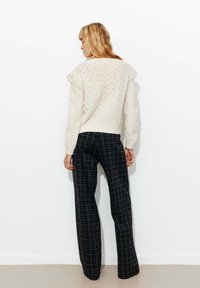 Pull en tricot crème avec un motif texturé et des épaules volantes, associé à un pantalon à carreaux sombres à jambes larges. Le tout sur un fond uni.