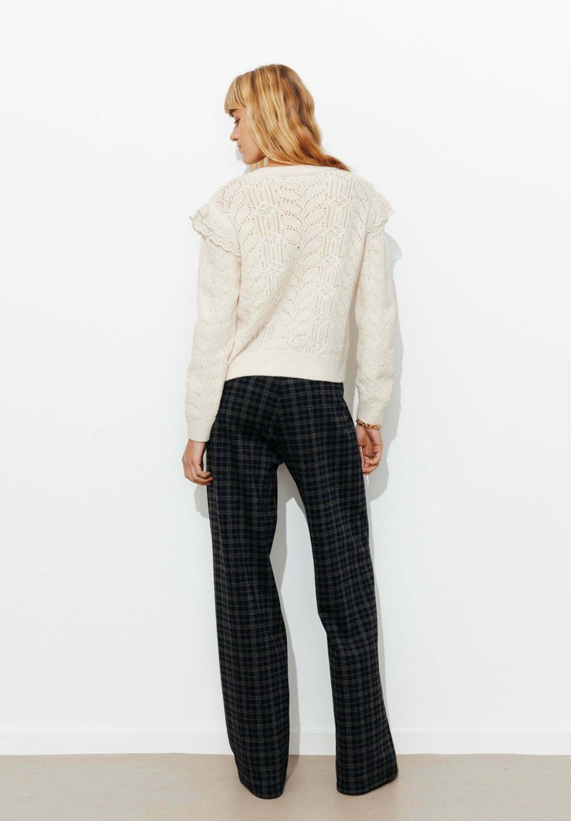 Pull en tricot crème avec un motif texturé et des épaules volantes, associé à un pantalon à carreaux sombres à jambes larges. Le tout sur un fond uni.