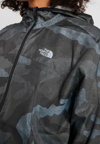 Chaqueta de camuflaje hecha de tela ligera, con tonos de negro y gris. Tiene capucha y cremallera frontal, con un logotipo en el pecho.