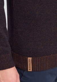 Granatowy sweter dzianinowy z teksturowanej tkaniny, z prążkowanym mankietem i dołem w kolorze brązowym. Na dole widoczne są metki z nazwą marki.