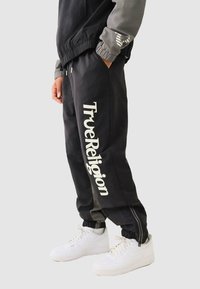Schwarze Jogginghosen mit weißem "TrueReligion"-Logo, elastischem Bund und Bündchen, seitlichen Reißverschlüssen und glatter Textur, kombiniert mit weißen Sneakers.