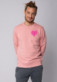 Sweatshirt rose clair avec un col rond, des manches longues et un grand cœur rose peint à la main sur la poitrine gauche. Texture de tissu lisse.