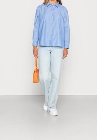 Chemise boutonnée rayée bleu clair, coupe décontractée, associée à un jean délavé et des baskets blanches. Tient un sac à main texturé orange.