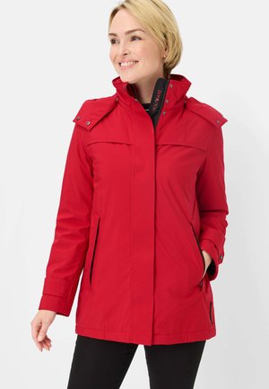Smiling woman wearing a red hooded jacket with zippered pockets and black pants, standing against a plain white background.
