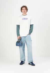 Modelis valkā baltu T-kreklu ar krāsainu Adidas logotipu, virsū uzvilkts zaļš garām piedurknēm. Zilās džinsās un tumši zila apavi pabeidz apģērbu.