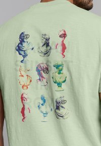 Lichtgroene t-shirt met een veelkleurige graphic van klassieke buste, sommige met een duikbril op, met een gedurfde achtergrondtekst.