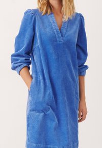Abito in velluto a coste blu con scollatura a V, maniche lunghe e polsini arrotolati. Presenta tasche laterali e una vestibilità comoda. Tessuto morbido.