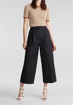 Femme portant un haut à manches courtes marron à motifs, un pantalon noir taille haute à jambes larges, et des sandales blanches à talons hauts, debout sur un fond gris.