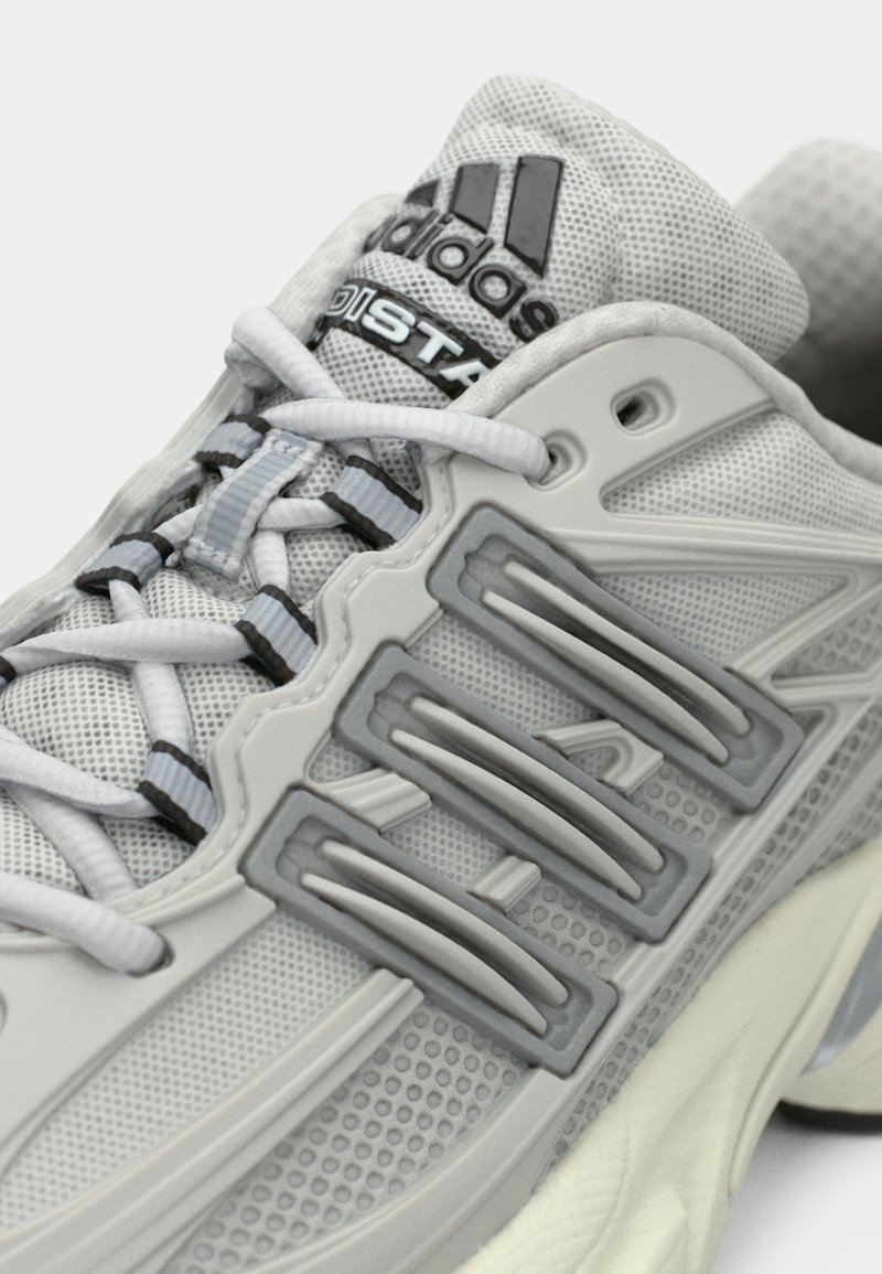 Scarpe da ginnastica grigie con tomaia in rete, suola in gomma, dettagli testurizzati e tre strisce distintive di un grigio più scuro sui lati.