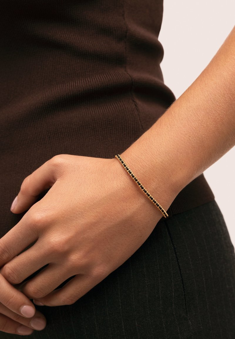 Gold bracelet with black stone accents, thin design, textured surface, worn on wrist, set against a dark brown fabric background.