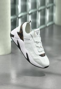 Scarpa sportiva bianca con elementi in mesh testurizzato e pelle, caratterizzata da un lato con un motivo nero e marrone, e un logo sulla linguetta e sul lato.