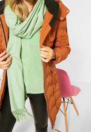 Vrouw draagt een oranje doorgestikte jas over een lichtgroene sjaal en top, staand bij een roze stoel met houten poten tegen een witte achtergrond.