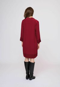 Burgundské midi šaty s dlhými rukávmi a hladkým povrchom. Majú vysoký golier a vrstvený lem. Nosí sa s čiernymi čižmami až po kolená.