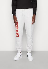 Ljust grå sweatpants med en röd grafik på vänster ben, elastiska muddar, dragsko i midjan, och ihopparade med vita Nike-sneakers.