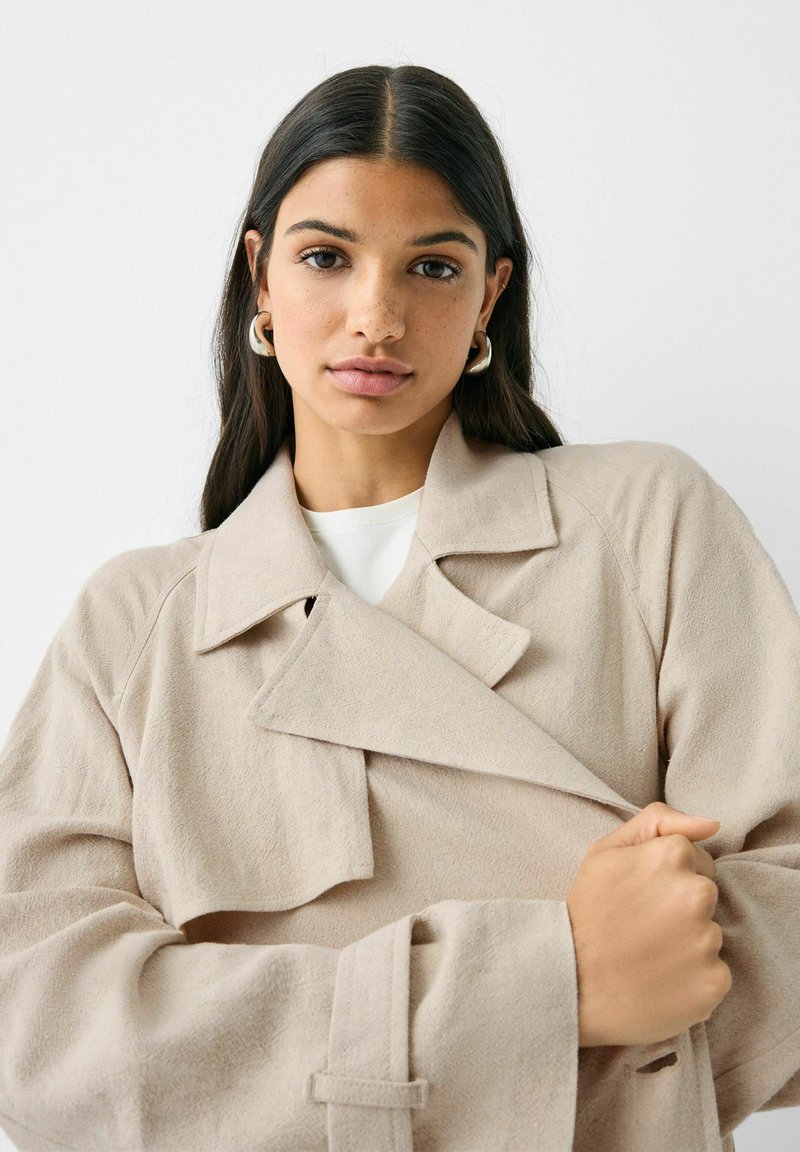 Cappotto trench oversized color beige con ampi rever e tessuto testurizzato. Presenta un design avvolgente con chiusura a cravatta e maniche ample.