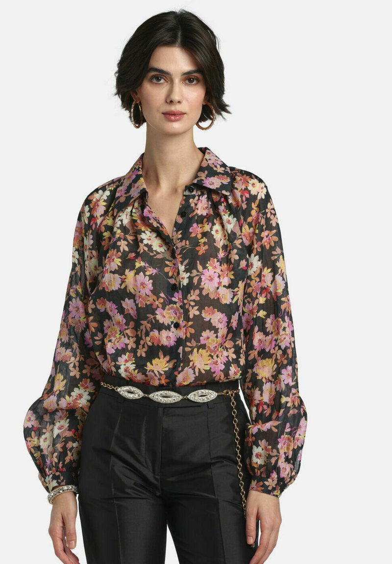 Donna che indossa una camicetta trasparente nera a fiori con maniche a palloncino, pantaloni neri a vita alta, una cintura decorativa e grandi orecchini a cerchio.