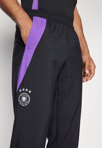 Zwarte sportbroek met paarse zijpanelen, elastische tailleband en geborduurd Duits voetbal-embleem met sterren en tekstdetail.