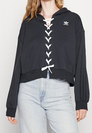 Vrouw draagt een zwarte Adidas cropped hoodie met witte veters aan de voorkant en lichtblauwe hoge taille jeans, staand tegen een effen achtergrond.