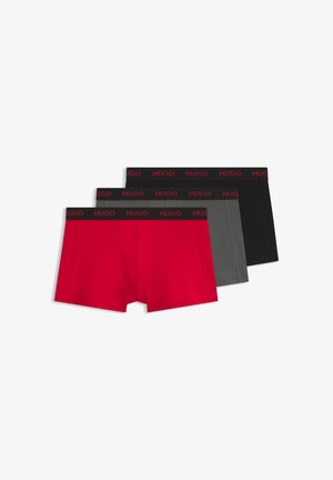 Jeune homme aux cheveux courts et foncés portant un boxer rouge avec une ceinture noire de la marque "Hugo", debout devant un fond blanc uni.