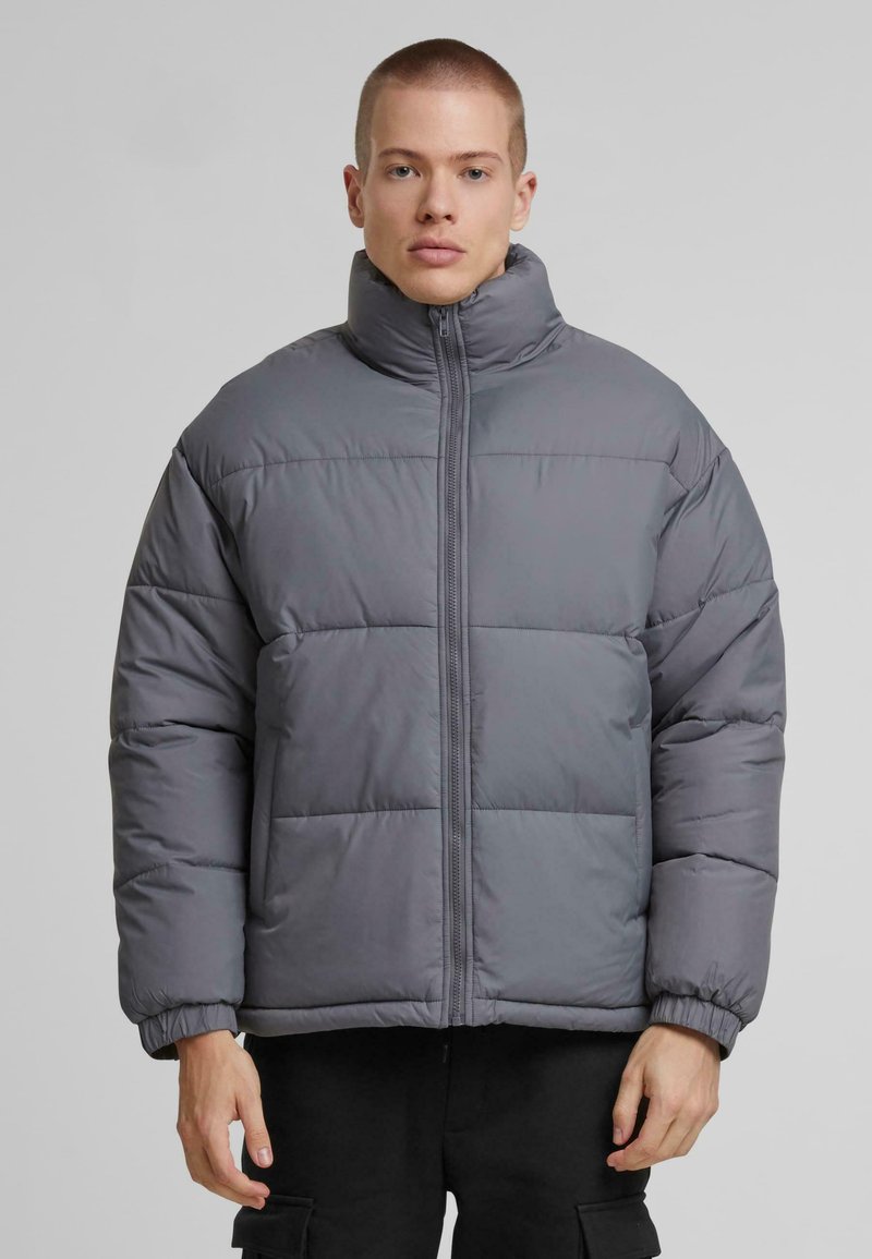 Grau gesteppte Daunenjacke mit hohem Kragen, ausgestattet mit einem Reißverschluss vorne und horizontalen Nähten. Weiche Textur und leicht übergroße Passform.