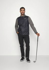 Temno modra vetrovka brez rokavov preko sive majice z dolgimi rokavi, črne hlače in črni golf čevlji. Drži golf palico, stoji ob preprostem ozadju.