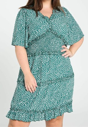 Femme portant une robe verte à pois blancs, taille froncée, bords volantés, manches courtes, main posée sur la hanche et la cuisse.